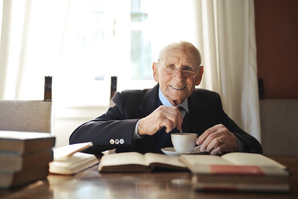 Senior man drinking hot beverage while reading book at table in light room